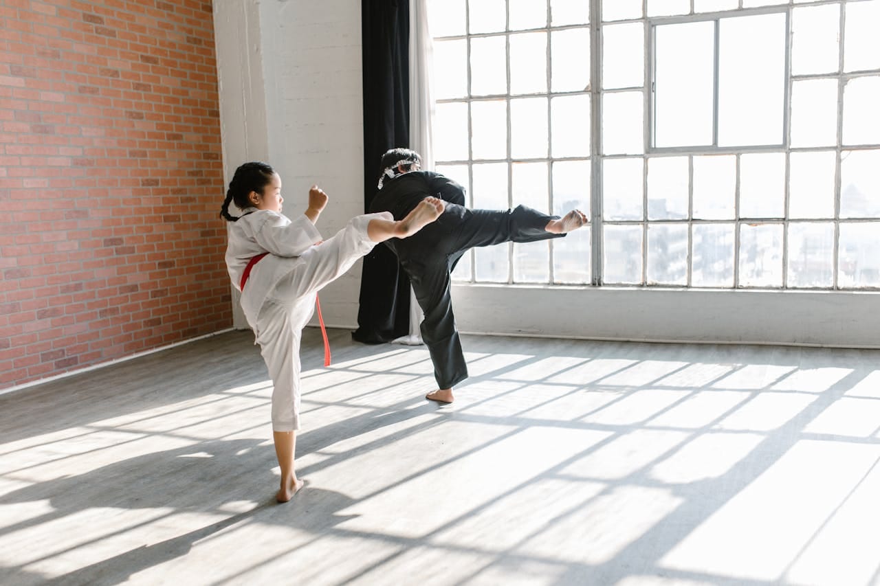 Two martial artists practicing high kicks in a brightly lit studio.